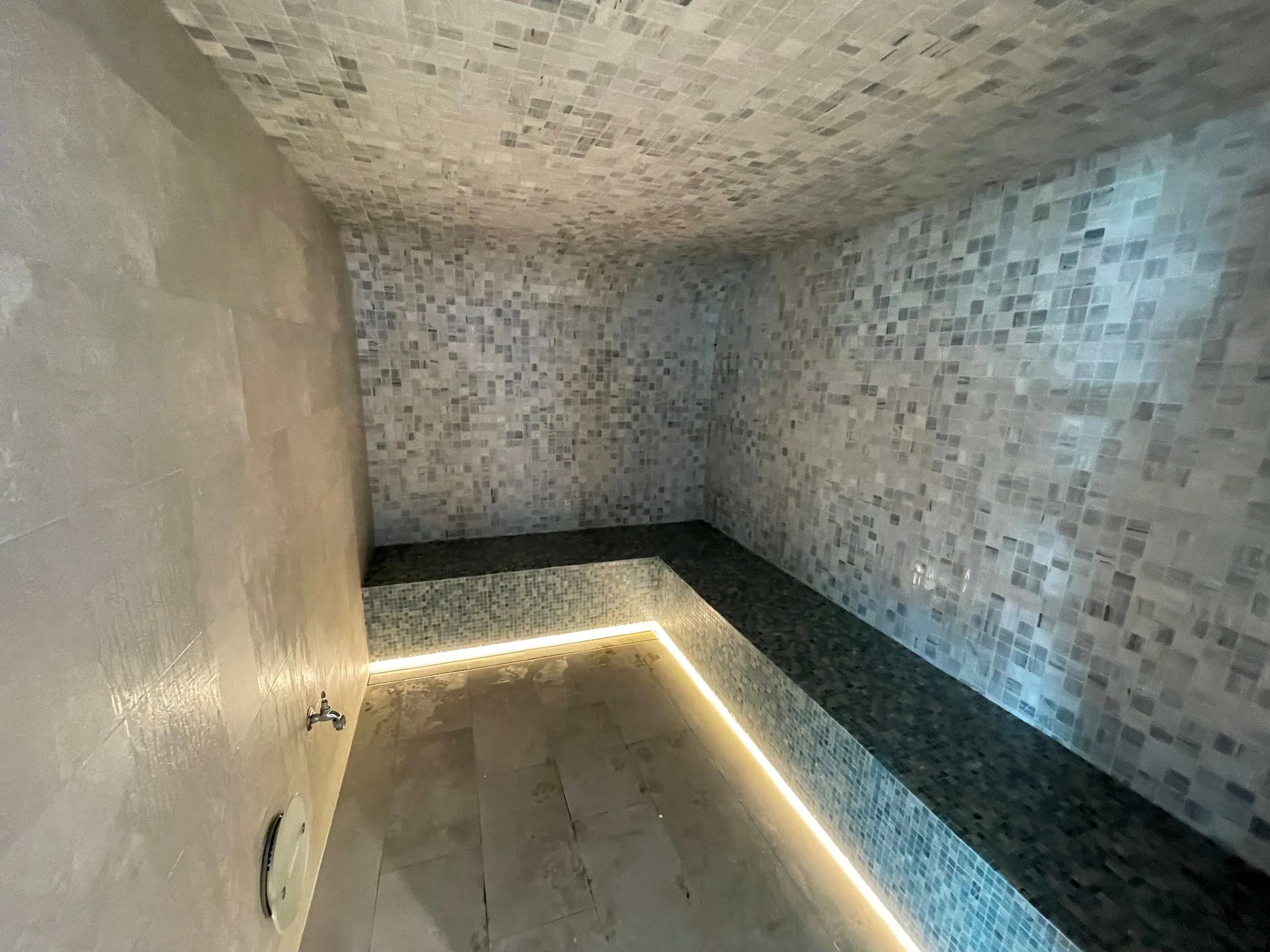 Barrel-vaulted steam room in mosaic tile with a stone bench outlined in soft linear light