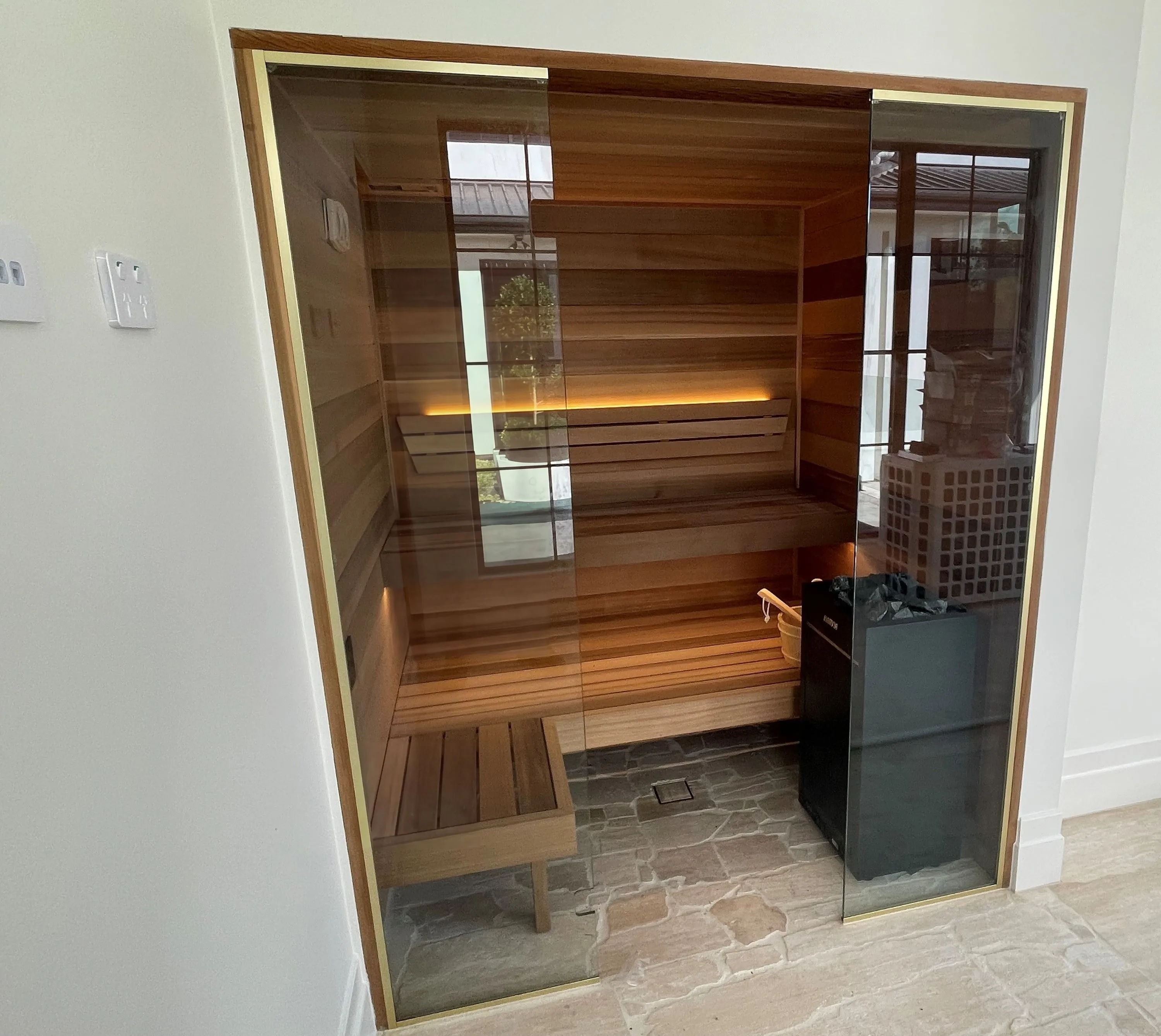 Gold-toned framed glass front of a horizontal timber home sauna with lit benches and a floor-standing heater