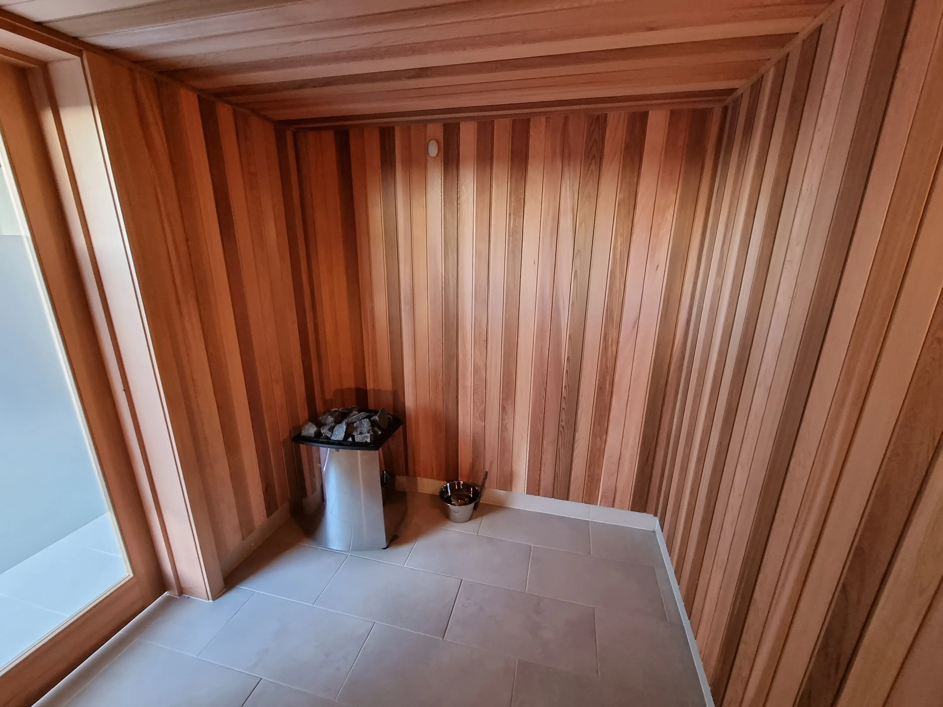 Floor-standing sauna heater beside a glazed side panel in a timber sauna