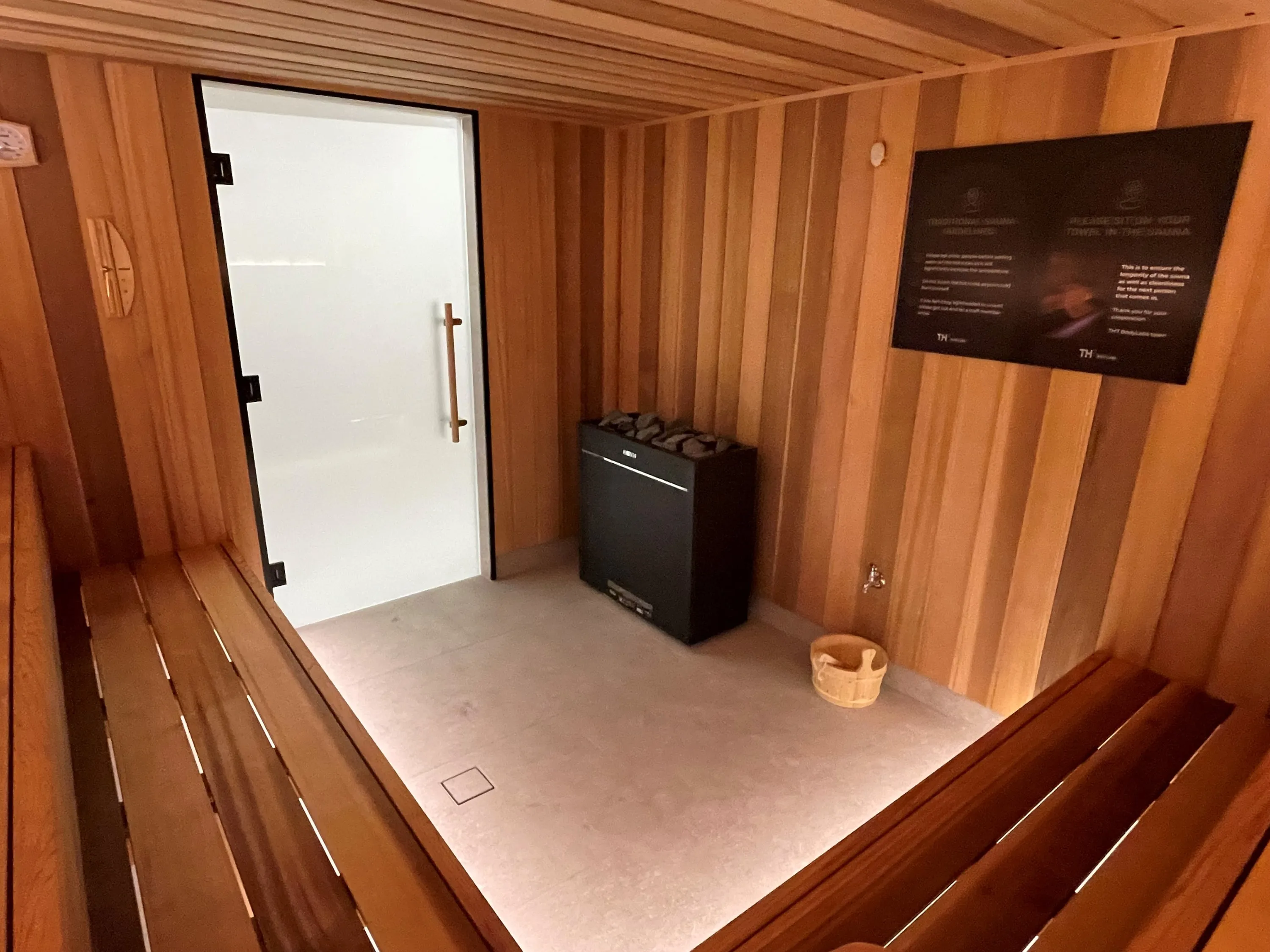 Floor-standing sauna heater with stones beside a frosted glass door and a guidelines board