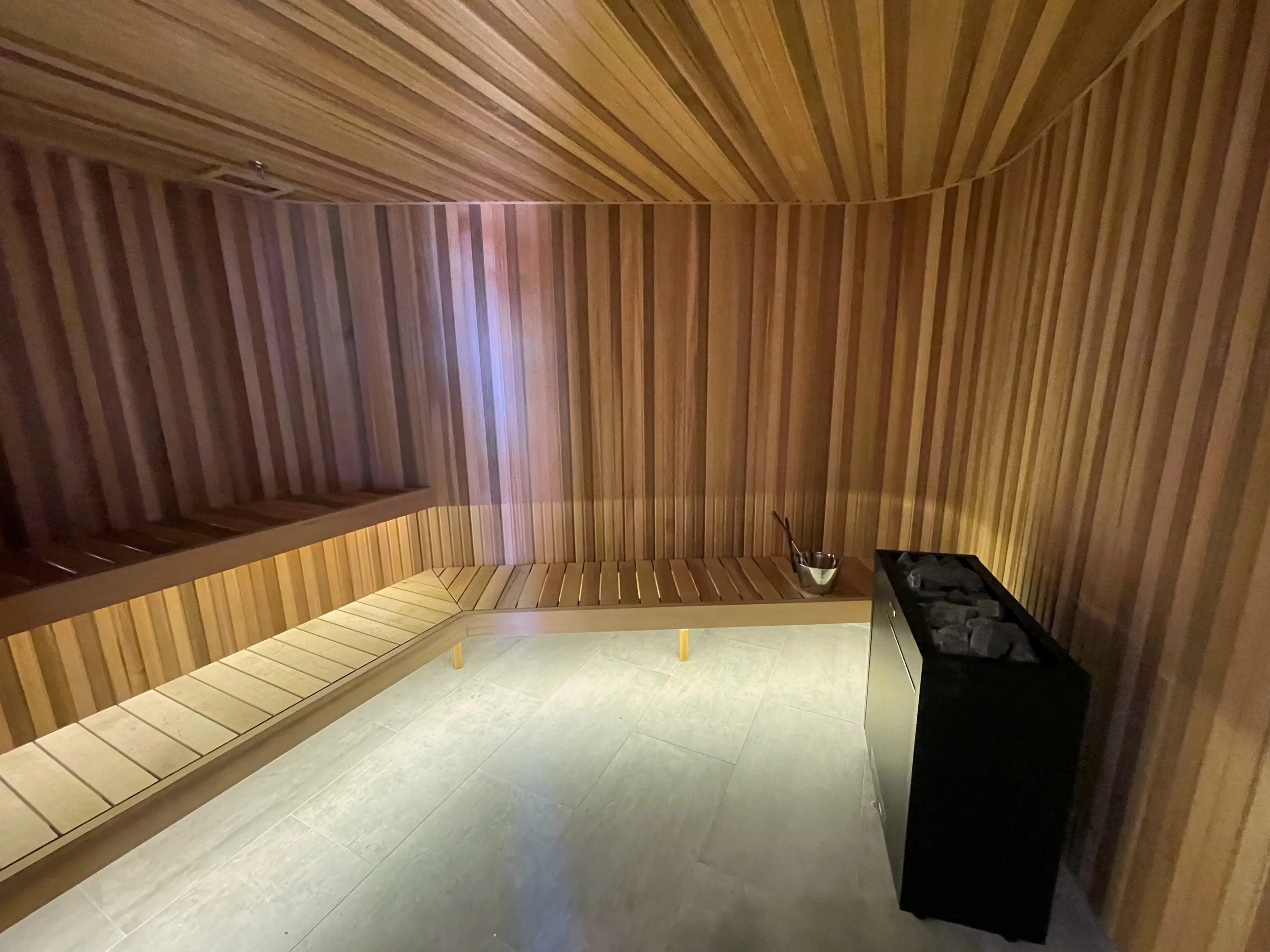 Lit timber benches wrapping the corner of the sauna beside a floor-standing heater with stones
