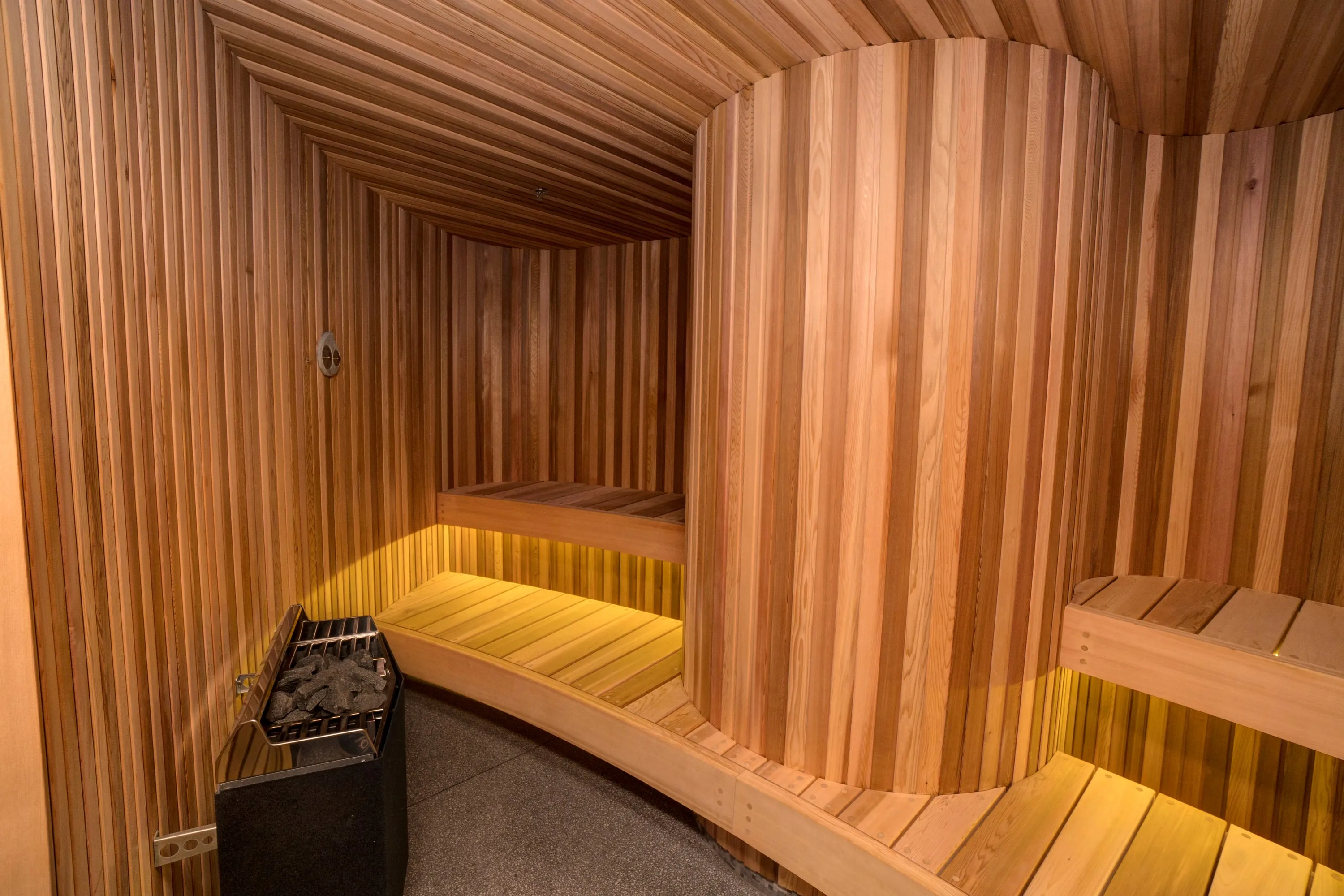 Curved cedar sauna with a lit bench following the wall and a heater filled with stones