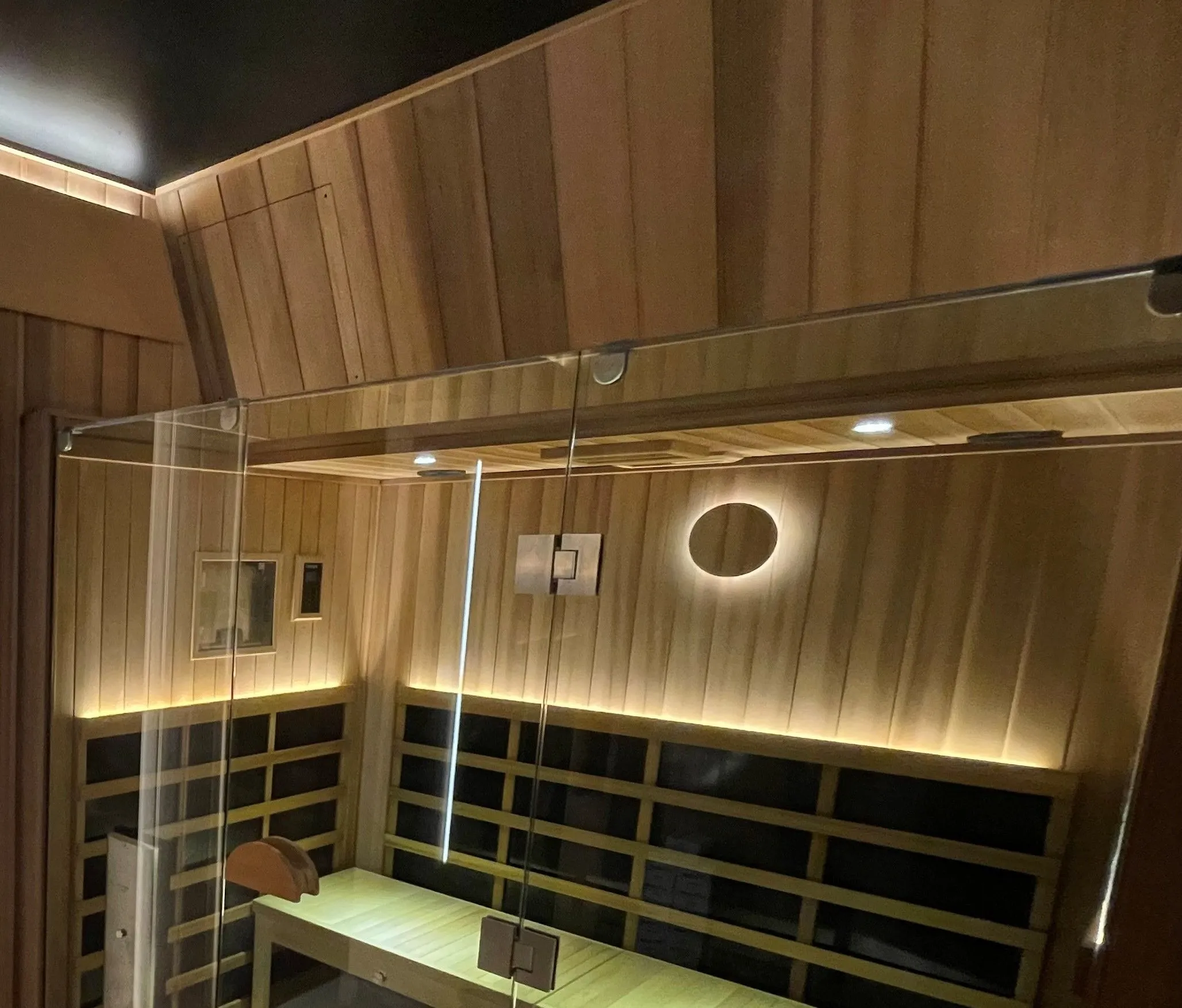 Glass-fronted infrared room with dark panels framed in timber along the backrest and a lit bench