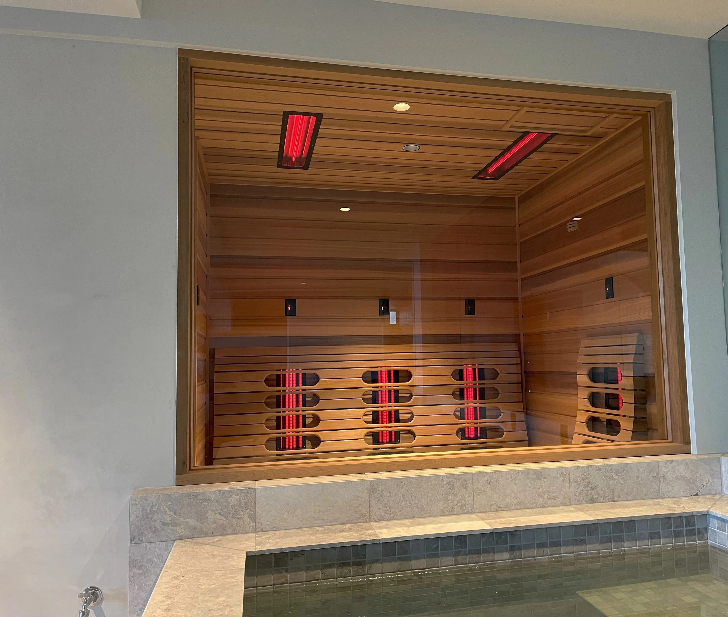 Timber infrared sauna seen through a large window above a pool, with red emitters glowing