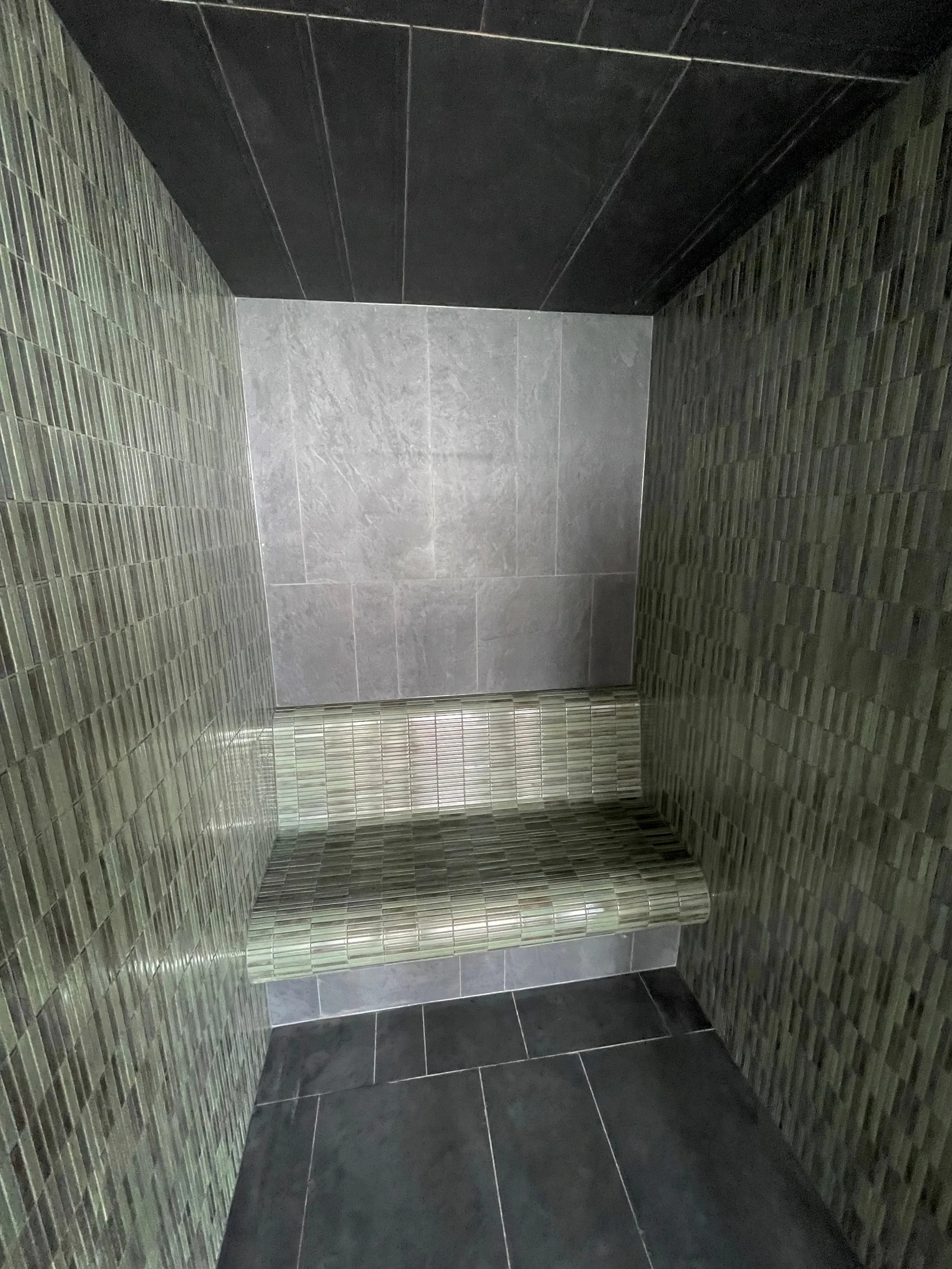 Narrow steam room in green mosaic tile with a tiled bench and a pale stone back wall