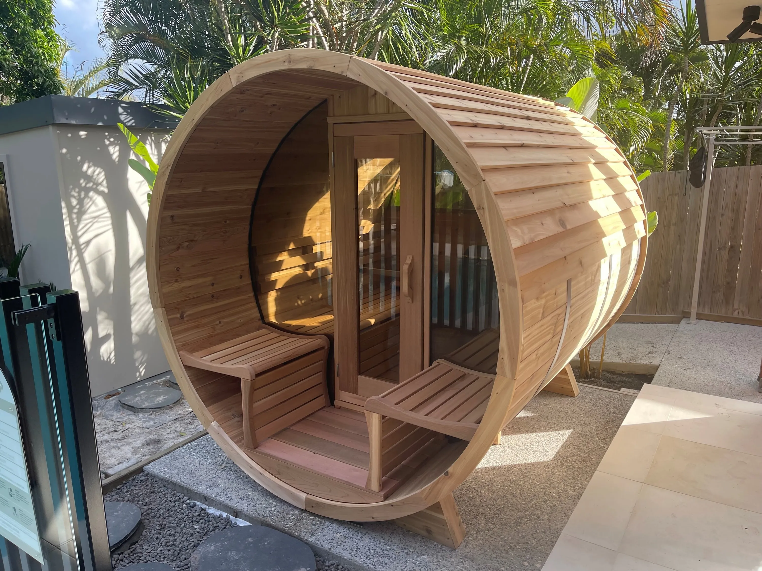 Timber barrel sauna with a covered front porch and benches in a tropical garden