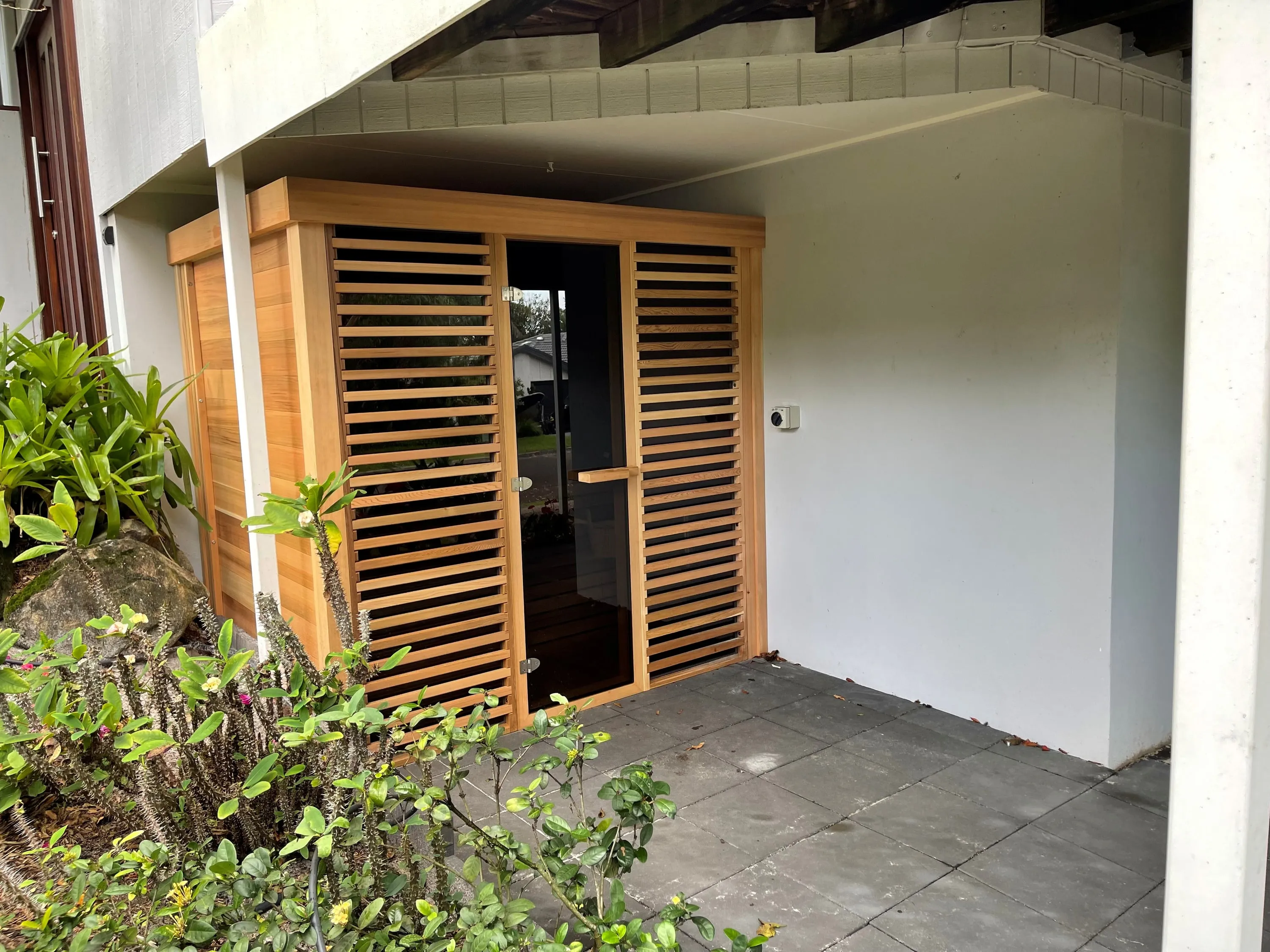 Pure Cube sauna tucked beneath a house beside a planted garden bed