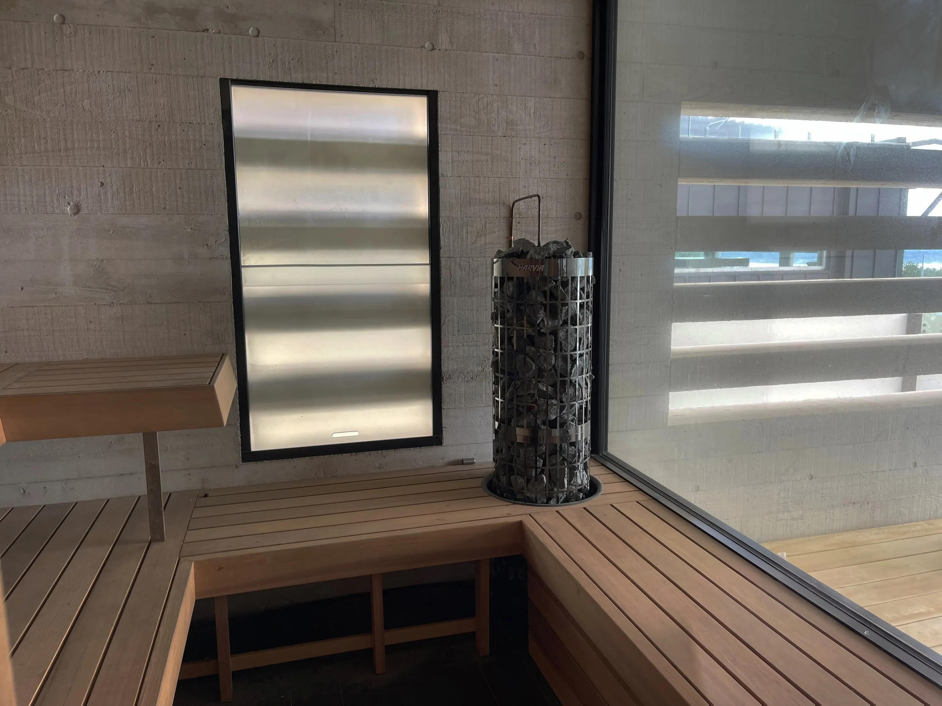 Tower heater beside a frosted window in board-formed concrete, timber benches and a full-height glass wall onto the deck