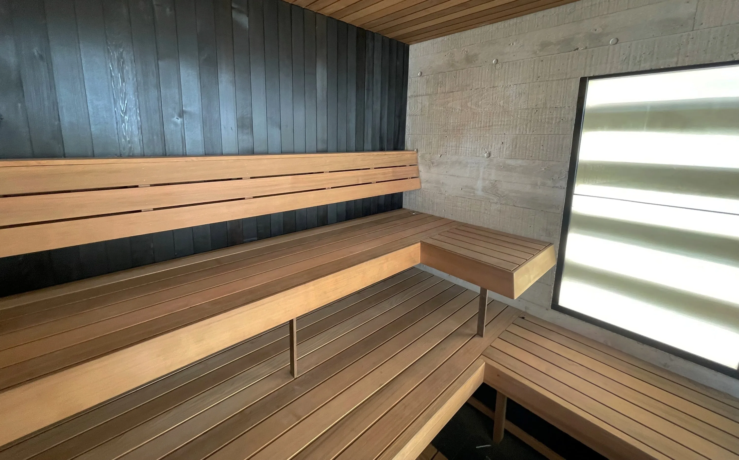 Two tiers of timber benches against a black timber wall and board-formed concrete, beside a glazed panel