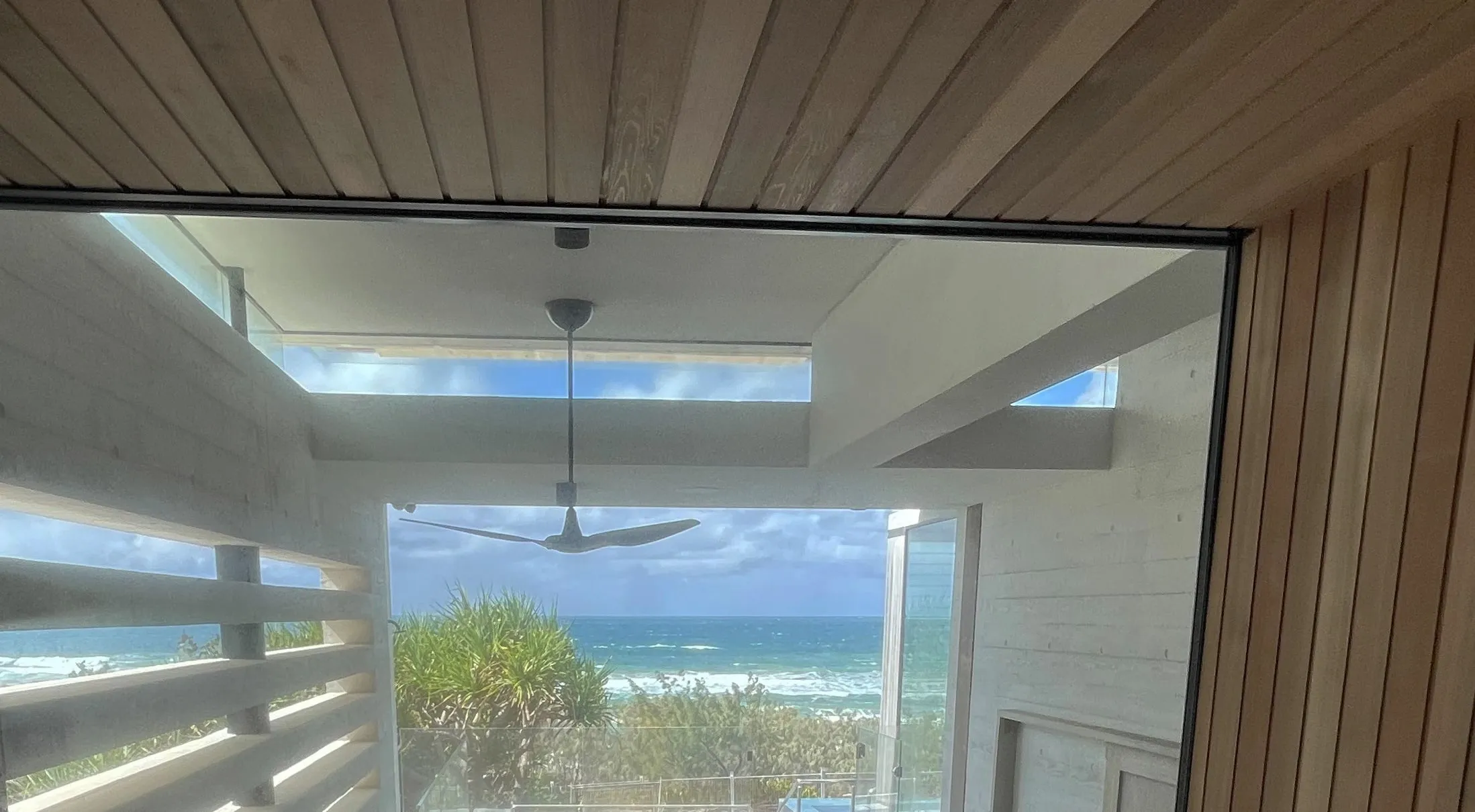 View from the sauna through a large window to concrete louvres, pandanus and the ocean beyond, under a timber ceiling