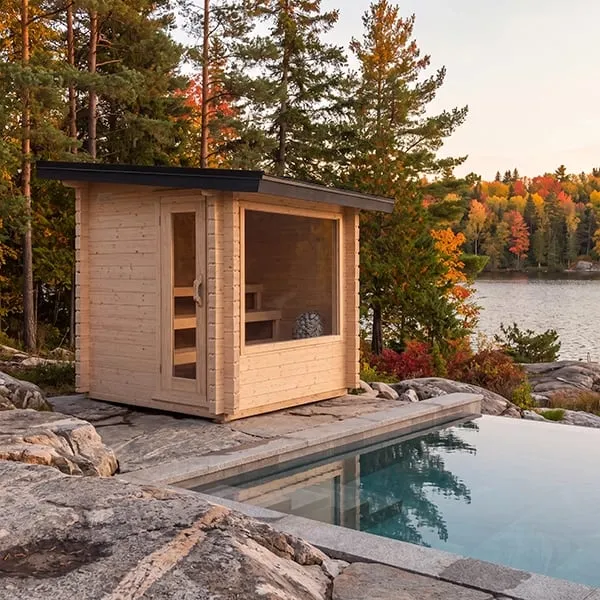 Manufacturer rendering: cabin sauna with light interlocking timber walls, flat dark roof, glass door and large front window, on rock beside a pool, lake and autumn forest.