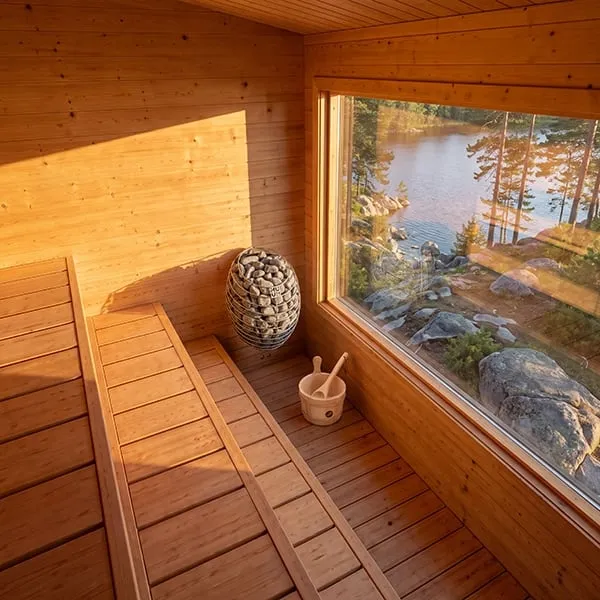 Manufacturer rendering: elevated view of a timber sauna interior with benches, caged electric heater and bucket, large window over a rocky lakeshore at sunset.