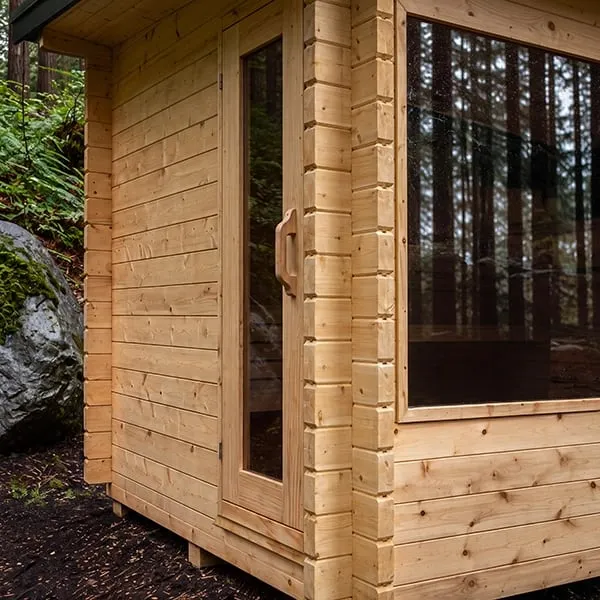 Corner detail of a light timber cabin sauna with interlocking corner joints, glass door with wooden handle and large window, in a forest beside a mossy boulder.