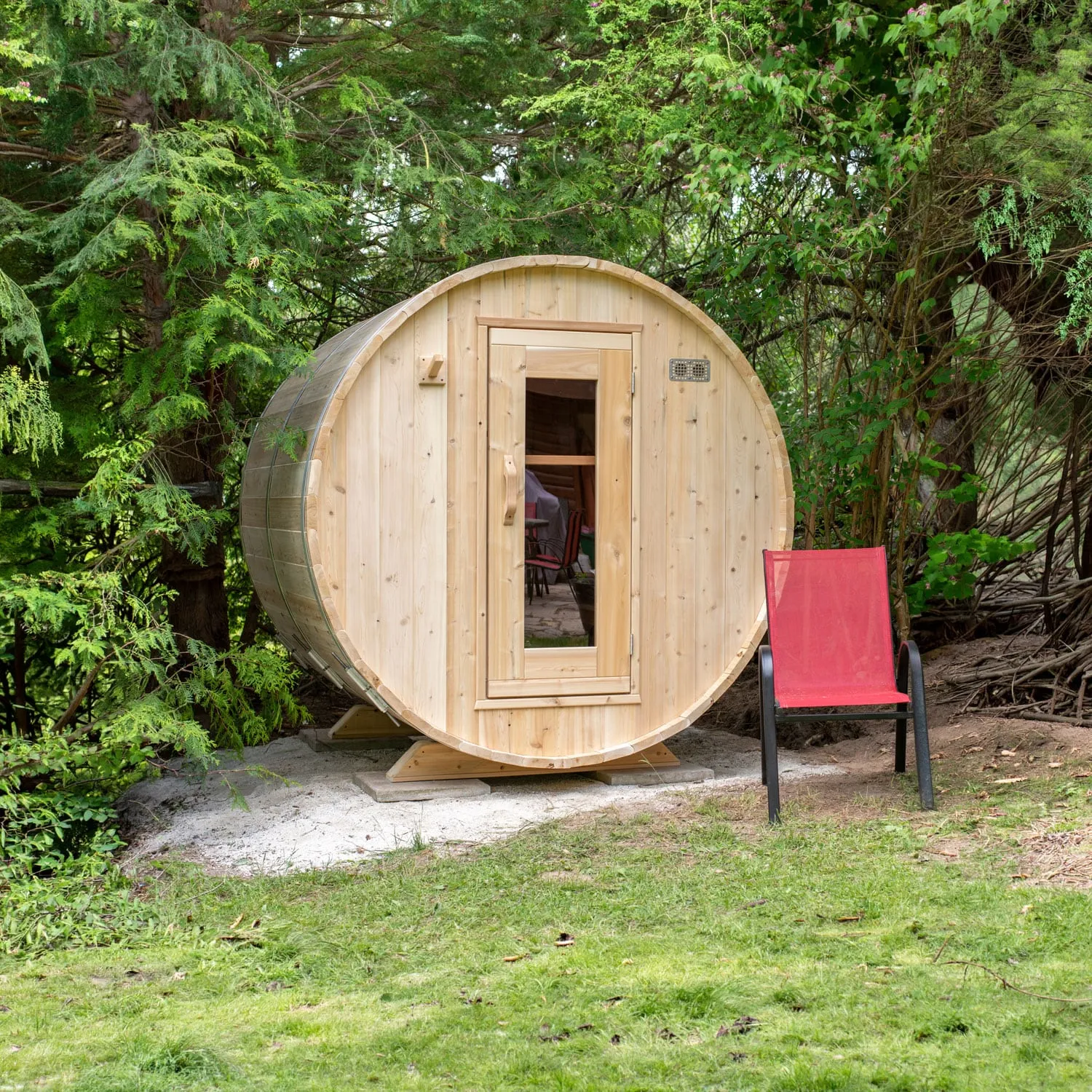 Barrel sauna in light knotty timber with metal bands and a glass-panel door, on concrete pavers among evergreen trees, red chair beside