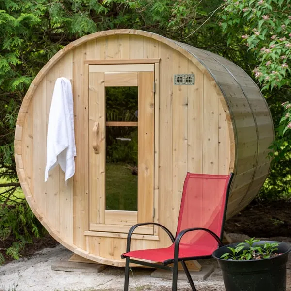 Front of a light timber barrel sauna with glass-panel door and towel on a hook, red chair and potted plant in a garden