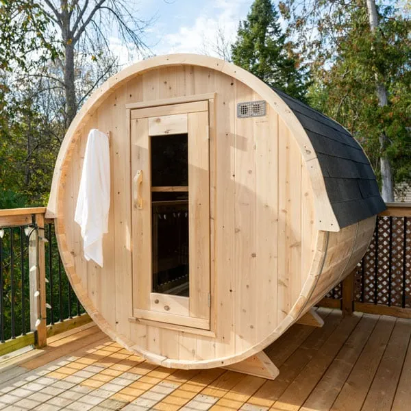 Light timber barrel sauna with dark shingle roof covering and glass-panel door, on a timber deck with trees behind