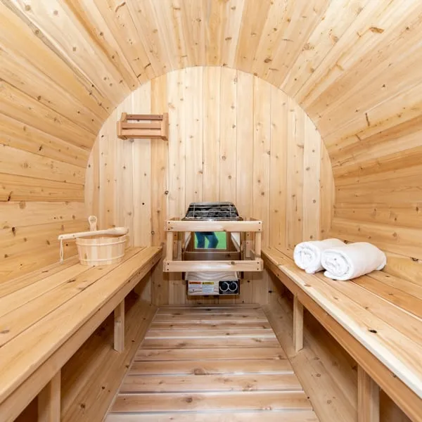 Barrel sauna interior with curved light knotty timber walls, benches on both sides, electric heater with stones in a timber guard, towels and bucket