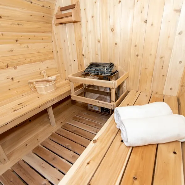 Inside a barrel sauna: electric heater with stones in a timber guard at the end wall, benches, rolled towels, bucket and slatted floor