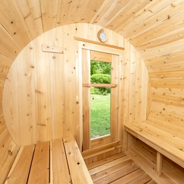 Barrel sauna interior looking toward the round end wall with glass-panel door onto a lawn, wall clock and timber benches