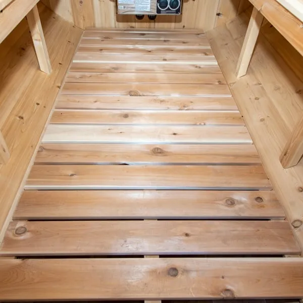 Slatted knotty timber floor inside a barrel sauna between bench supports, heater controls at the far end