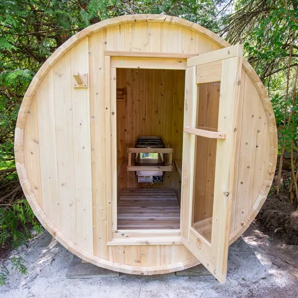 Light timber barrel sauna with door open showing an electric heater in a timber guard at the back, on pavers among trees