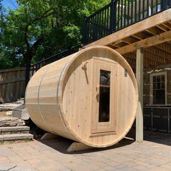 Light timber barrel sauna with metal bands and glass-panel door, on brick paving beneath an elevated house deck