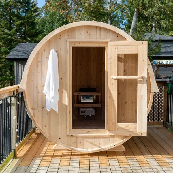 Light timber barrel sauna on a timber deck with door open showing heater and benches, towel hanging on the front