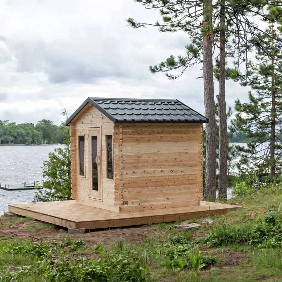 Small log-style cabin sauna in light knotty timber with interlocking corners, black metal tile gable roof, glass door and two narrow windows, on deck by a lake