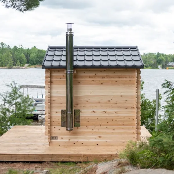 Rear wall of log-style cabin sauna with stainless chimney flue and heat shield, black metal tile roof, on a timber deck by a lake