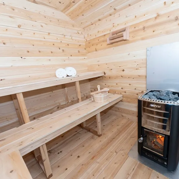 Cabin sauna interior with wood-burning heater with glass firebox door and stones, metal heat shield, two-tier benches, towels and bucket