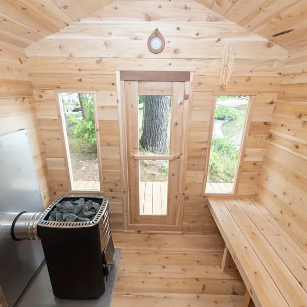 Cabin sauna interior with wood-burning heater and heat shield, glass-panel door between two narrow windows, peaked ceiling and wall clock