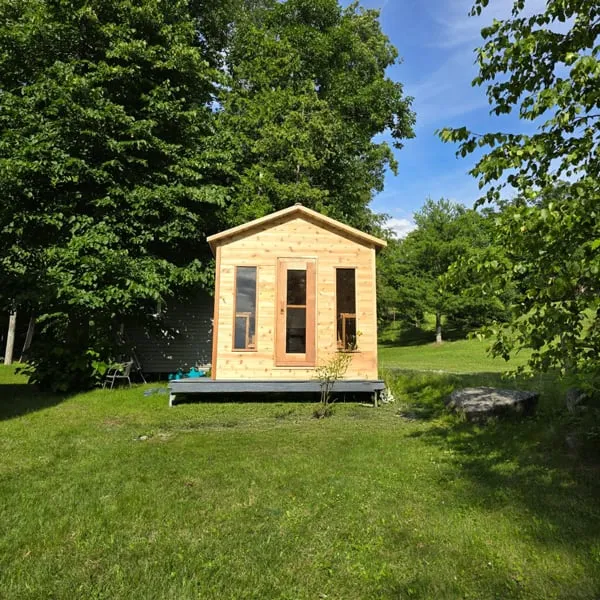 Gable-roofed cabin sauna in light timber with glass-panel door and two narrow windows, on a low dark deck in a grassy yard with trees
