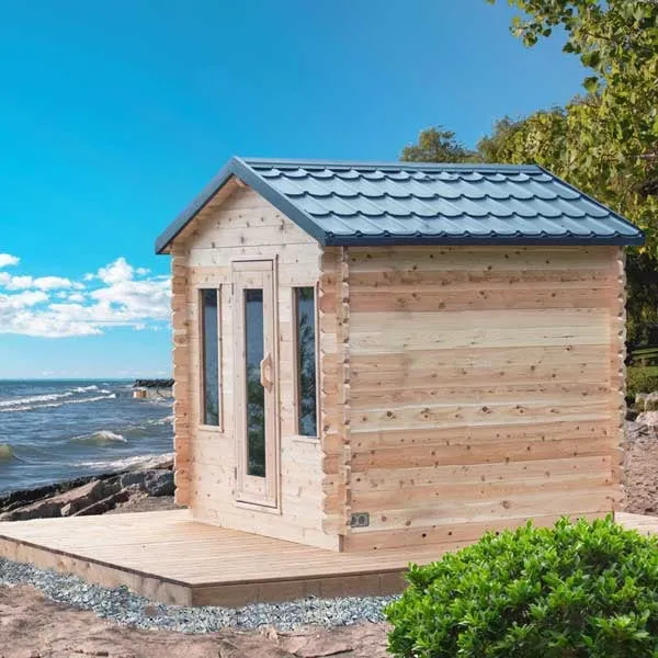 Log-style cabin sauna with black metal tile gable roof, glass door and two narrow windows, on a timber deck at a lakeshore