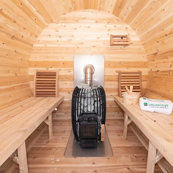 Barrel sauna interior in knotty cedar: benches along both sides with backrests, black caged wood-burning stove with stones and flue against a steel heat shield.