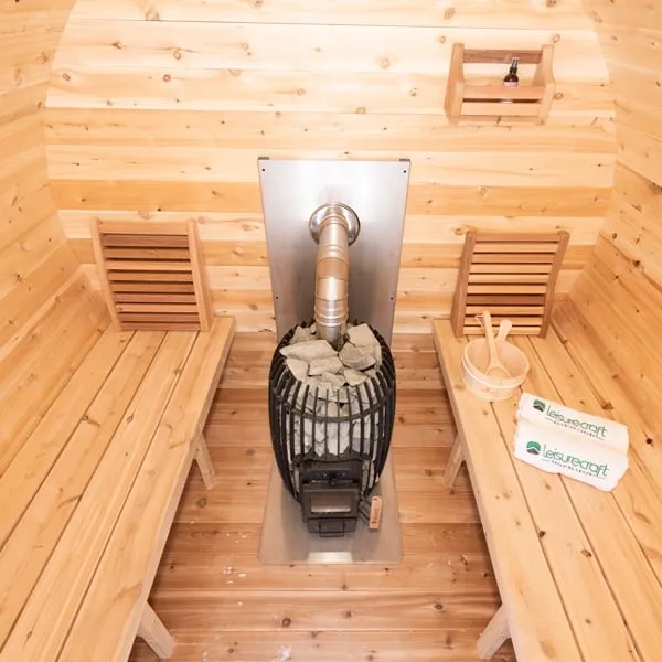 Elevated view inside a cedar barrel sauna: wood-burning stove with stones and stainless flue on a steel heat shield between two side benches, bucket, ladle and towels.