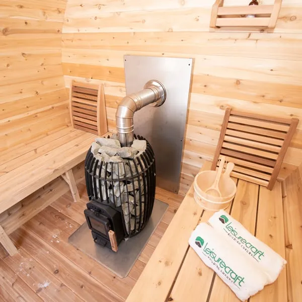 Angled close view of a caged wood-burning sauna stove with stones, stainless flue and steel heat shield, beside cedar benches with backrest, bucket, ladle and rolled towels.