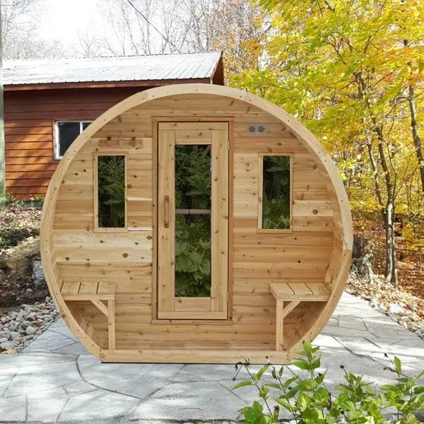 Front view of a cedar barrel sauna with porch benches, glass-panel door and two windows, on stone paving in autumn woodland in front of a cabin.