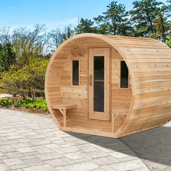 Three-quarter view of a light cedar barrel sauna with recessed porch, benches, glass-panel door and two windows, on a paver patio with spring trees.