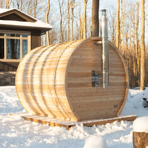 Rear three-quarter view of a cedar barrel sauna on a timber base in snow, stainless chimney flue rising through the rear end wall, house and bare trees behind.