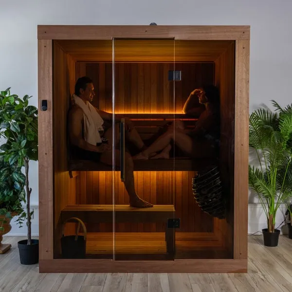 Indoor sauna cabinet in reddish-brown timber with full-height glass front, two people seated on two-tier benches, warm LED strip lighting, caged heater, potted plants.