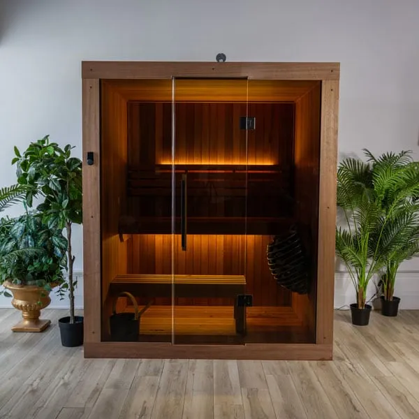 Front view of an indoor sauna in reddish-brown timber with full-height glass front and door, two-tier benches, LED strip lighting and caged heater, in a white room.