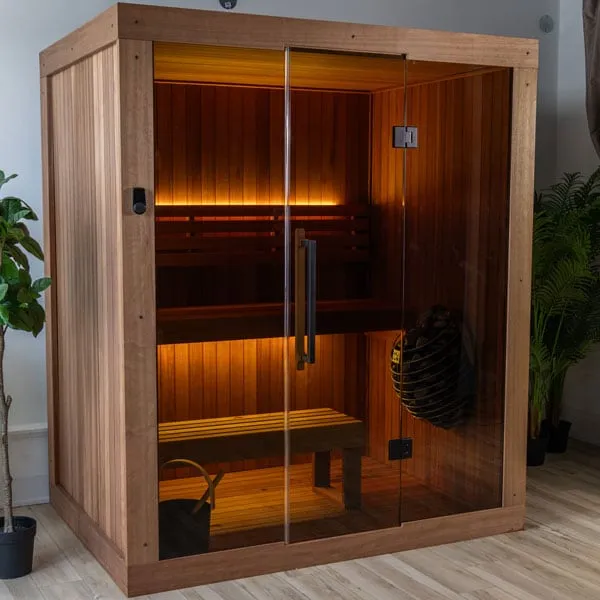 Three-quarter view of an indoor sauna with vertical reddish-brown timber side panel, full-height glass front with door, lit benches and caged heater.