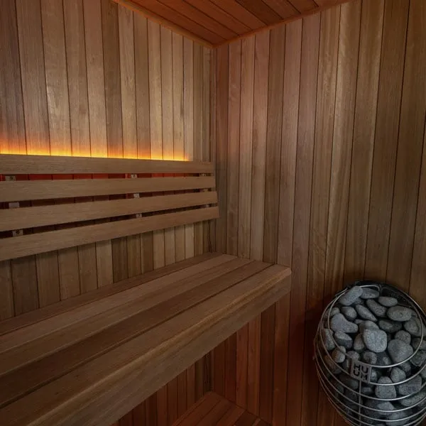 Indoor sauna interior with vertical reddish-brown timber lining, bench with slatted backrest, LED strip lighting and caged electric heater with stones.