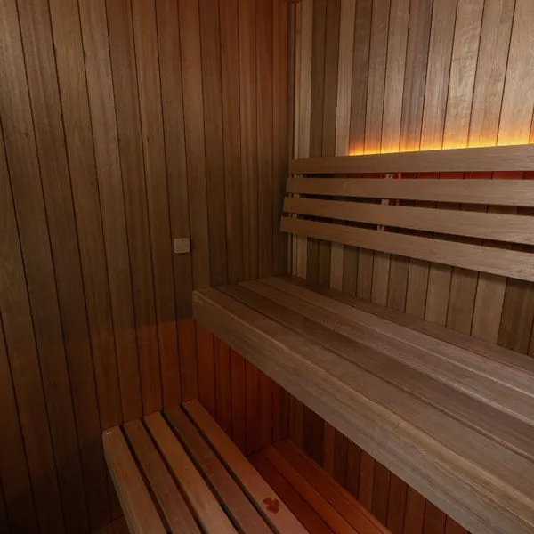 Corner of an indoor sauna interior in vertical reddish-brown timber with upper bench, slatted backrest, concealed LED strip and lower step bench.