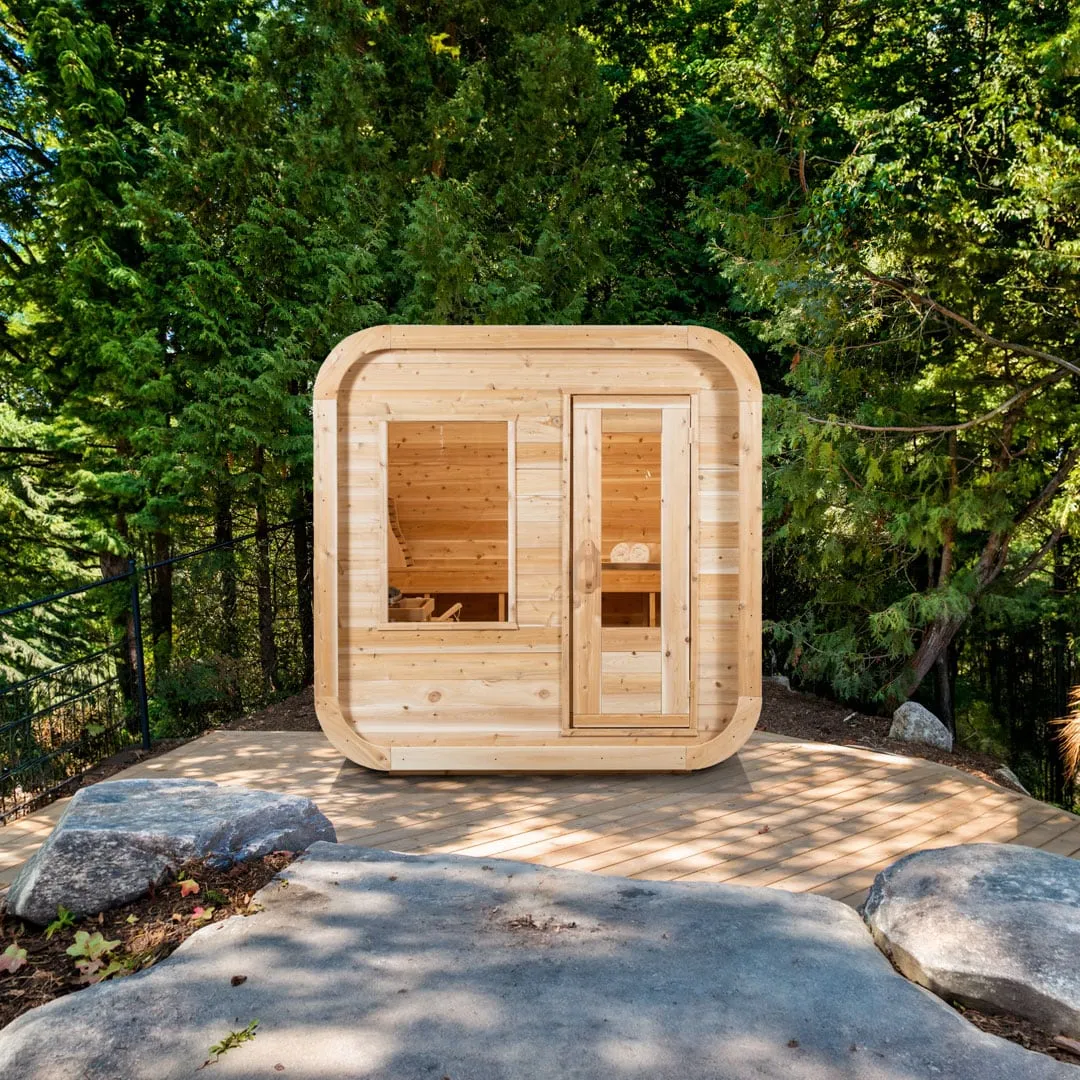 Front of a square sauna with rounded corners in light knotty cedar, large front window and glass-panel door, on a timber deck among evergreen trees.