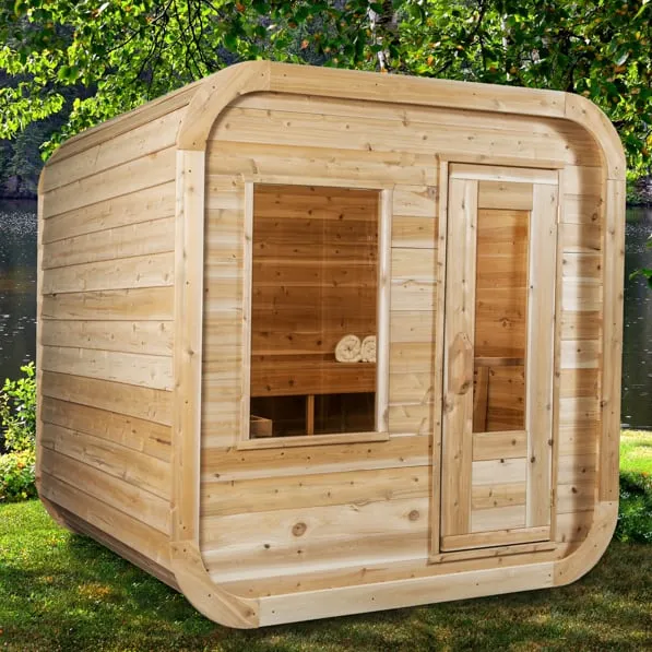 Three-quarter view of a rounded-corner cube sauna in light knotty cedar with front window and glass-panel door, on grass beside a lake under trees.