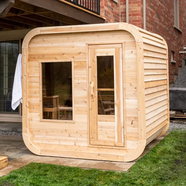 Rounded-corner cube sauna in light cedar with front window and glass-panel door, on a paver patio beside a brick house, towel hanging on the side.
