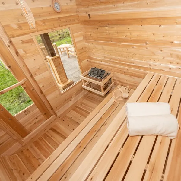 Elevated interior view of a cedar cube sauna: electric heater with stones in a timber guard, front window to a patio, benches, bucket, ladle, towels and thermometer.
