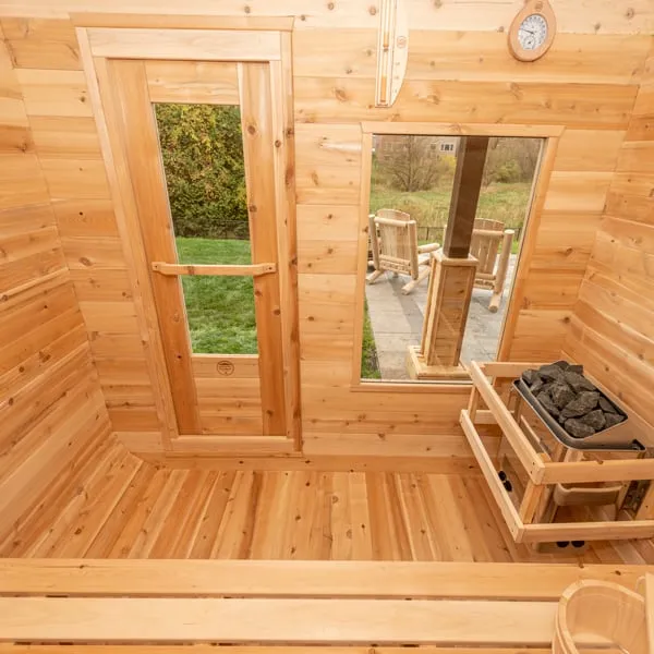 Cedar cube sauna interior looking toward the glass-panel door and front window, electric heater with stones in a timber guard, wall thermometer.
