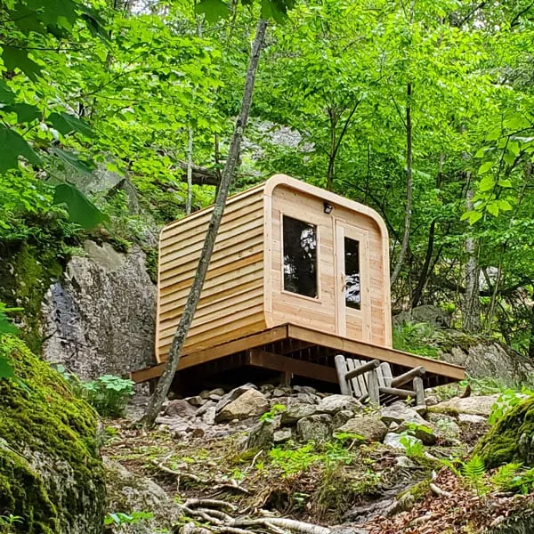Rounded-corner cedar cube sauna with front window and glass door on a raised timber platform on a rocky forest hillside, log bench beside.